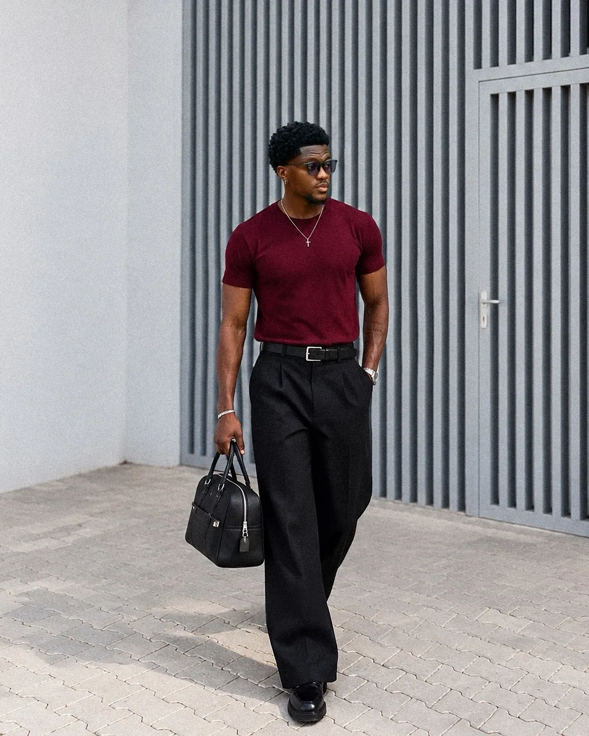 Model walking in a burgundy tee and pleated black trousers, carrying a leather bag